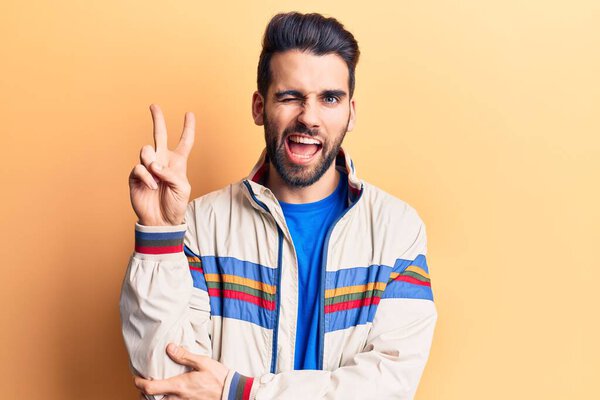 Young handsome man with beard wearing casual jacket smiling with happy face winking at the camera doing victory sign. number two. 