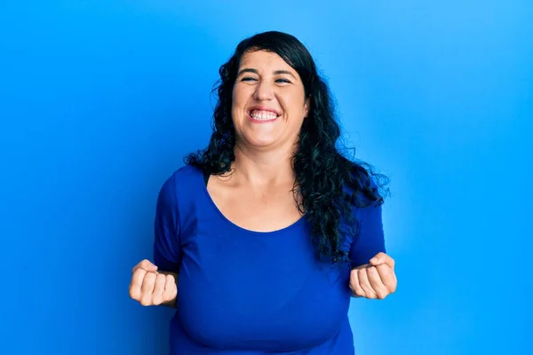 プラスサイズのブルネットの女性は カジュアルブルーのシャツを着て非常に満足し 腕を上げて 笑顔と成功のために叫んで勝者のジェスチャーを行う興奮しています お祝いのコンセプト — ストック写真