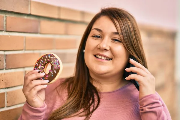 在城市里 他用智能手机和吃甜甜圈的年轻的恐慌加身材的女人 — 图库照片