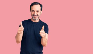 Orta yaşlı yakışıklı bir adam günlük tişört giyer. Başarı işareti yapar. Elle olumlu jest yapar, gülümseyerek mutlu olur. Neşeli ifade ve kazanan jest. 