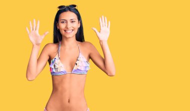 Bikini ve güneş gözlüğü takan genç ve güzel Latin kız on numarayı gösterip işaret ederken kendinden emin ve mutlu bir şekilde gülümsüyor.. 