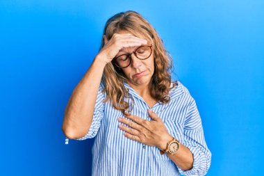 Orta yaşlı sarışın kadın günlük kıyafetler giyiyor ve gözlük takıyor hastalık, ateş, grip ve soğuk algınlığı için alnına dokunuyor. 