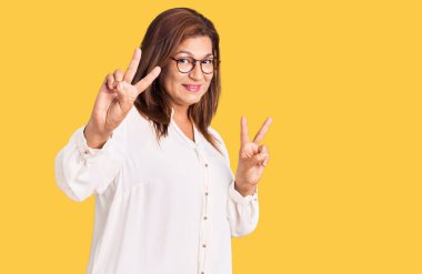 Orta yaşlı beyaz kadın, günlük kıyafetler giyiyor, gülümsüyor parmaklarını göstererek zafer işareti yapıyor ve gözlerini kaldırıyor.