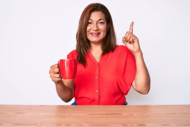 Orta yaşlı esmer kadın masada oturmuş kahve içiyor. Bir fikir ya da soru işareti ile şaşırmış, mutlu yüzlü, bir numara. 