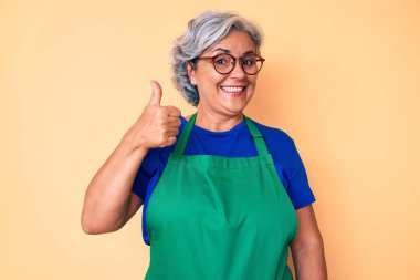 Önlük ve gözlük takan kıdemli İspanyol kadın mutlu ve pozitif gülümsüyor, başparmağıyla mükemmel ve onay işareti yapıyor. 