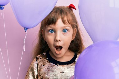 Little caucasian girl kid holding balloons afraid and shocked with surprise and amazed expression, fear and excited face. 