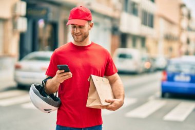 Beyaz atlı genç teslimatçı akıllı telefon kullanıyor ve şehirde kağıt torba taşıyor..