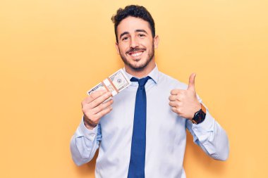 İş kıyafetleri giymiş genç İspanyol adam elinde dolarlarla mutlu ve pozitif gülümsüyor, başparmağıyla mükemmel ve onay işareti yapıyor. 