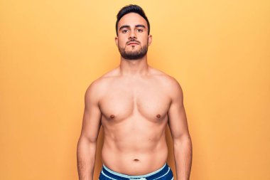 Sakallı, kolsuz tişörtlü, sarı arka planda duran yakışıklı bir genç. Yüzünde ciddi bir ifade var. Basit ve doğal bir şekilde kameraya bakıyorsun..