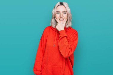 Sıradan kıyafetler giyen genç sarışın kız kameraya kendinden emin bakıyor kollarını kavuşturup gülümsüyor ve çenesini kaldırıyor. Olumlu düşünmek. 