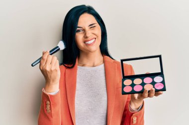 Makyaj fırçası tutan beyaz bir kadın ve kırmızı bir palet, kameraya seksi bir ifadeyle bakıyor, neşeli ve mutlu bir yüz.. 