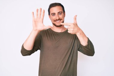 Genç ve yakışıklı bir adam gündelik giysiler giyer ve yedinci parmaklarını göstererek gülümserken kendinden emin ve mutlu olur.. 