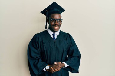 Yakışıklı siyahi adam mezuniyet şapkası ve tören cübbesi takıyor ve ellerini kavuşturup gülümsüyor. Başarı ve iyimserlik 