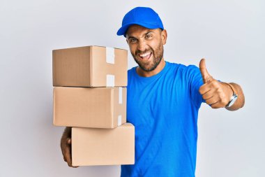 Sakallı, kurye üniforması giyen, teslimat paketleri taşıyan, elinde olumlu bir jest yapmayı onaylayan, gülümseyen ve başarı için mutlu bir adam. kazanan jest. 