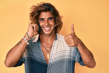 Genç İspanyol adam akıllı telefondan konuşuyor mutlu ve olumlu gülümsüyor, başparmağıyla mükemmel ve onay işareti yapıyor. 