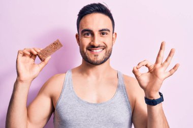 Sakallı, sakallı, enerji proteini yiyen, pembe arka planda yaşayan yakışıklı bir adam parmaklarıyla işaret veriyor, gülümseyerek mükemmel bir sembol oluşturuyor.