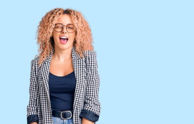 Kıvırcık saçlı, iş ceketli ve gözlüklü genç bir kadın kameraya seksi bir ifadeyle bakıyor, neşeli ve mutlu bir yüzü var.. 