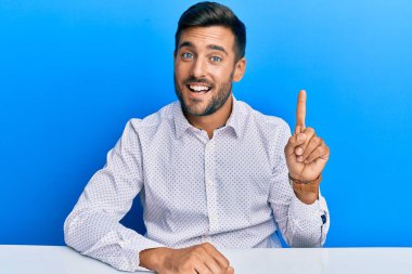 İş kıyafetleri giymiş yakışıklı İspanyol adam masaya oturmuş başarılı bir fikirle işaret ediyor. Heyecanlı ve mutlu. Bir numara.. 