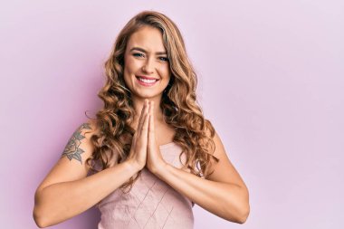 Genç sarışın kız gündelik giysiler içinde ellerini birleştirip dua ediyor af diliyor kendinden emin bir şekilde gülümsüyor.. 