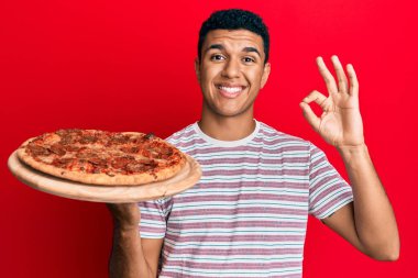 Young arab man holding italian pizza doing ok sign with fingers, smiling friendly gesturing excellent symbol 