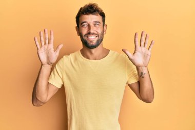 Sarı arka planda sarı tişört giyen sakallı yakışıklı bir adam on numarayı gösterirken kendinden emin ve mutlu bir şekilde gülümsüyor.. 