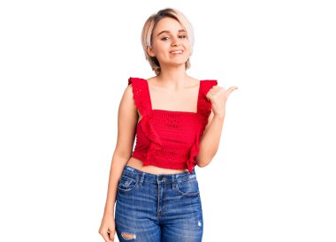 Genç, güzel, sarışın, sıradan kıyafetler giyen, mutlu yüzlü, gülümseyen, başparmağıyla yan tarafı gösteren bir kadın.. 