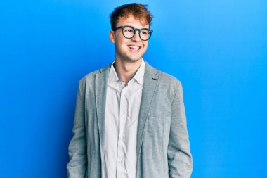 Zarif kıyafetler giyen ve gözlüklü genç beyaz adam yüzünde gülümsemeyle yüzünü yana doğru çeviriyor, doğal bir ifade. Kendinden emin gülen. 