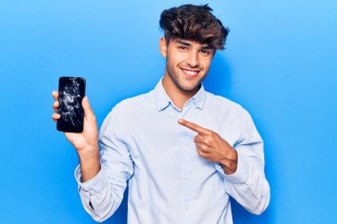 Kırık akıllı telefon tutan genç İspanyol adam kırık ekranı gösteriyor. Mutluluktan eli ve parmağı ile işaret ediyor. 