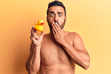 Genç İspanyol adam elinde ördek oyuncağı tutarak ağzını eliyle kapatıyor, şokta ve hata yapmaktan korkuyor. şaşırmış ifade 