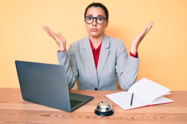 Otel resepsiyonunda çalışan esmer bir kadın. Laptop kullanıyor. Kolları açık ve kafası karışık.. 