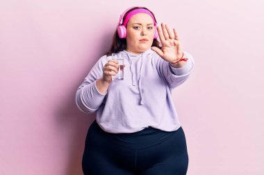 Genç ve cüsseli bir kadın bir bardak su içiyor kulaklık takıyor, kulaklık takıyor, dur işareti yapıyor ciddi ve kendinden emin bir ifadeyle savunma hareketi yapıyor. 