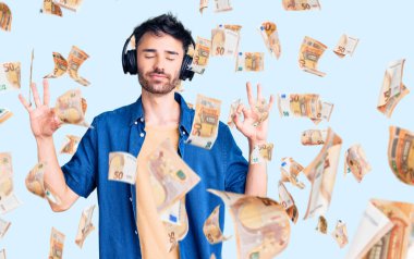 Kulaklık kullanarak müzik dinleyen, gözleri kapalı gülümseyen genç İspanyol adam parmaklarıyla meditasyon hareketi yapıyor. yoga konsepti.