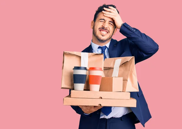 Young hispanic man wearing suit holding takeaway coffee and food stressed and frustrated with hand on head, surprised and angry face