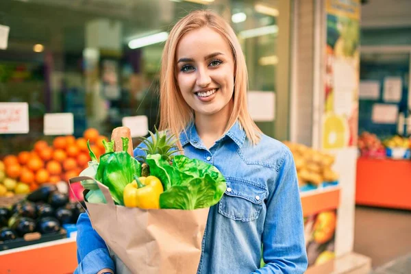 年轻的金发姑娘笑着高高兴兴地拿着食品杂货纸袋站在超市前 — 图库照片