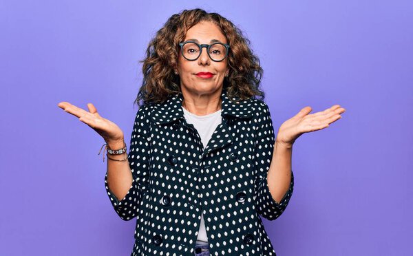 Middle age beautiful woman wearing casual coat and glasses over isolated purple background clueless and confused with open arms, no idea and doubtful face.