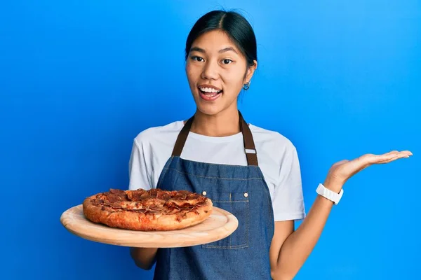 若いです中国人女性身に着けていますウェイターエプロン保持イタリアのピザの達成を祝います幸せな笑顔と勝者式とともに上げ手 — ストック写真