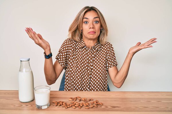 Beautiful caucasian woman drinking healthy almond milk clueless and confused with open arms, no idea and doubtful face. 
