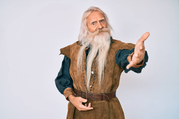 Old senior man with grey hair and long beard wearing viking traditional costume smiling friendly offering handshake as greeting and welcoming. successful business. 