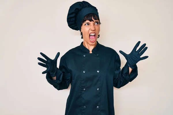 Beautiful brunettte woman wearing professional cook uniform crazy and mad shouting and yelling with aggressive expression and arms raised. frustration concept.