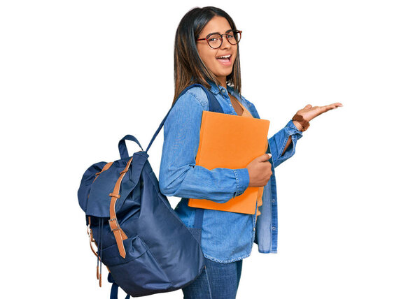 Young latin girl wearing student backpack and holding books pointing aside with hands open palms showing copy space, presenting advertisement smiling excited happy 