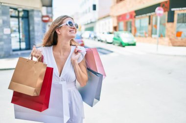 Genç sarışın kadın gülümsüyor. Şehrin caddesinde alışveriş torbalarını tutarken mutlu.