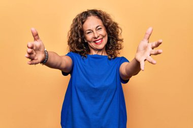 Orta yaşlı güzel bir kadın, günlük tişört giyip izole edilmiş sarı arka planda dikiliyor ve kameraya sarılıp gülümsüyor. Mutluluğu kucaklayan neşeli bir ifade..