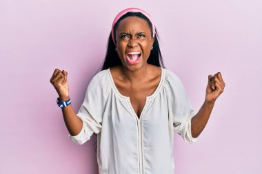 Pembe arka plan üzerine günlük kıyafetler giyen genç Afrikalı bir kadın çılgın ve kızgın bir şekilde bağırıyor ve agresif bir ifade ve kollarla bağırıyor. hüsran kavramı. 