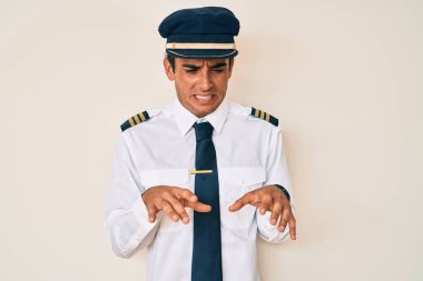 Young hispanic man wearing airplane pilot uniform disgusted expression, displeased and fearful doing disgust face because aversion reaction. 