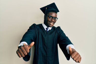 Mezuniyet şapkası ve tören cübbesi giyen yakışıklı siyahi adam elleriyle olumlu jest yapmayı onaylıyor, gülümseyerek ve başarı için seviniyor. kazanan jest. 