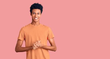 Genç Afro-Amerikan adam, günlük kıyafetler giyip ellerini birleştiriyor ve gülümsüyor. Rahatlamış ve neşeli. Başarı ve iyimserlik 