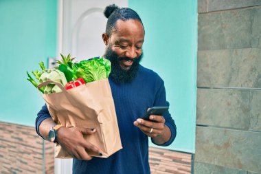 Sakallı Afrikalı Amerikalı adam elinde süpermarketten alınmış kağıt torbalarla akıllı telefon kullanıyor.