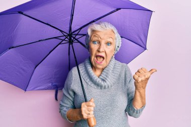 Yaşlı, gri saçlı, elinde mor şemsiyeyle baş parmağını kaldırmış, ağzı açık gülümseyen bir kadın. 
