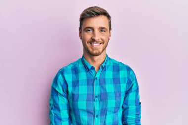 Yakışıklı beyaz bir adam günlük kıyafetler giyiyor. Pozitif ve mutlu görünüyor. Kendine güvenen bir gülümsemeyle dişlerini gösteriyor. 