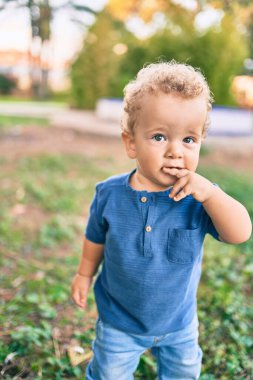 Üzgün küçük çocuk, güneşli bir günde parkta diş etlerine dokunduğu için parmaklarını dudaklarına koyuyor. Güzel sarı saçlı erkek bebek yeni bebek dişleri için acı çekiyor.